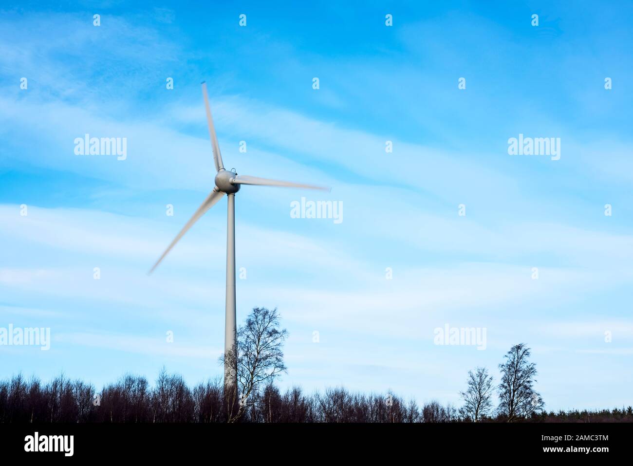 Three bladed wind turbine generator and pylon situated above Stracathro, Angus, Scotland, UK surrounded by trees, blades blurred revealing motion. Stock Photo