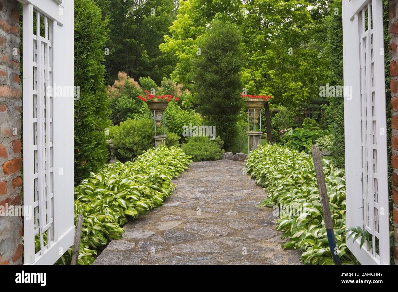 Red brick columns with white wooden opened fence gate and flagstone path bordered by Hosta plants in private backyard formal garden in early summer. Stock Photo