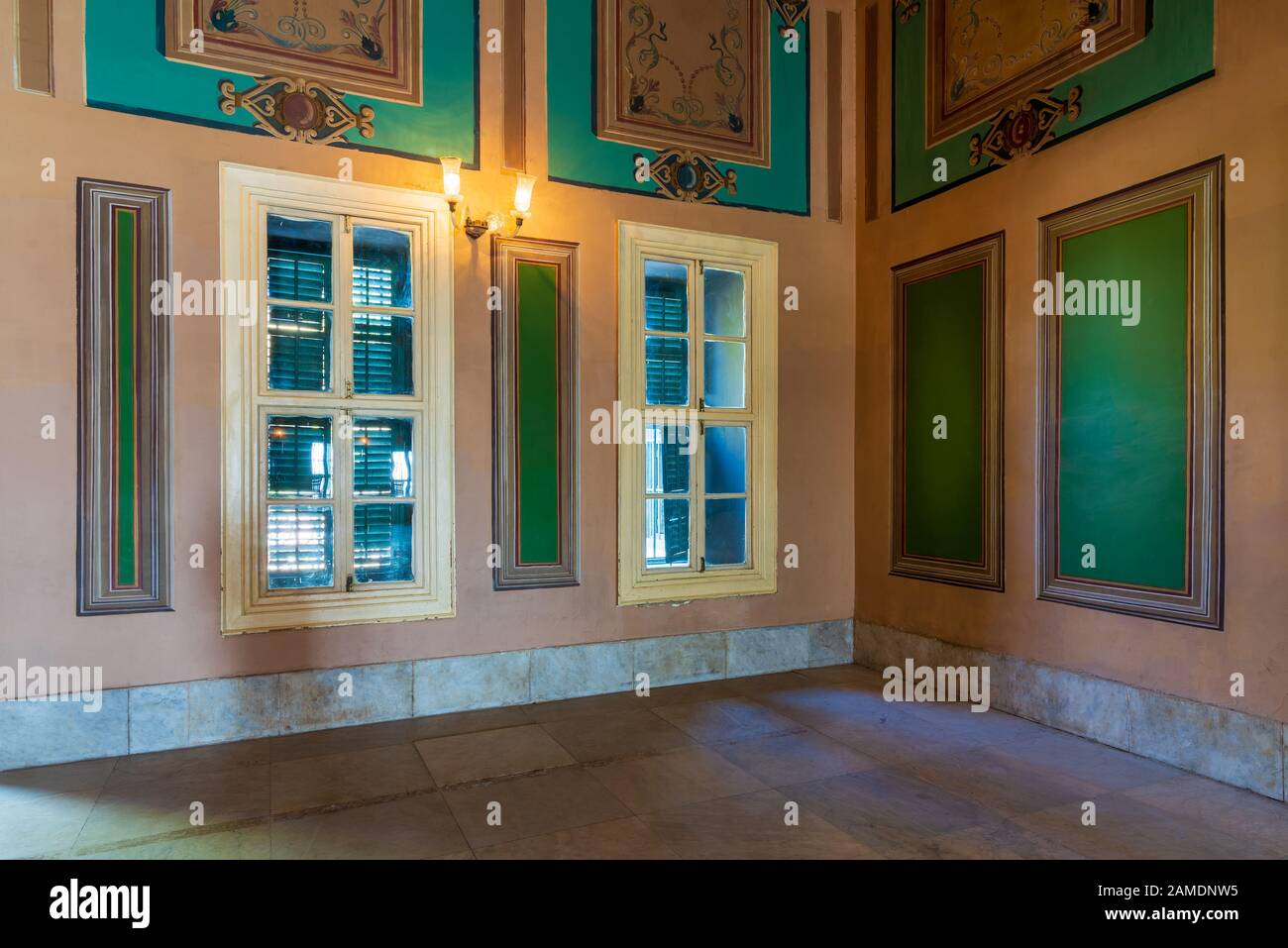 Corner orange wall with two wooden windows with green shutters, beautiful elegant rectangular green frames with ornate border, and white tiled marble floor Stock Photo