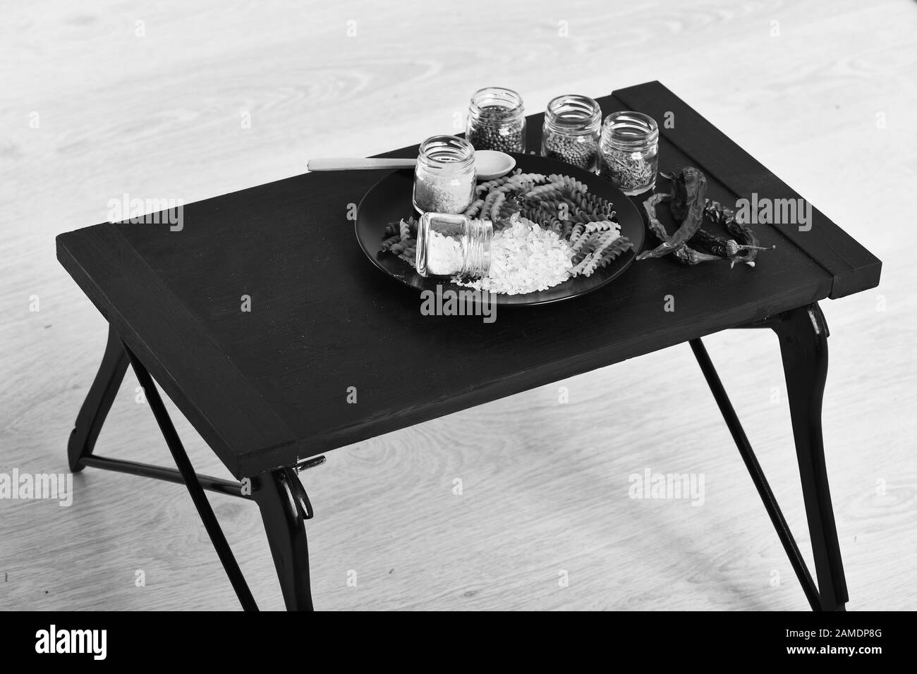 Taste and spicy cuisine concept. Jars of spices near plate of pasta on grey wooden background. Italian dish placed on wooden table tray. Fusilli with salt on plate, spoon and chili pepper nearby. Stock Photo