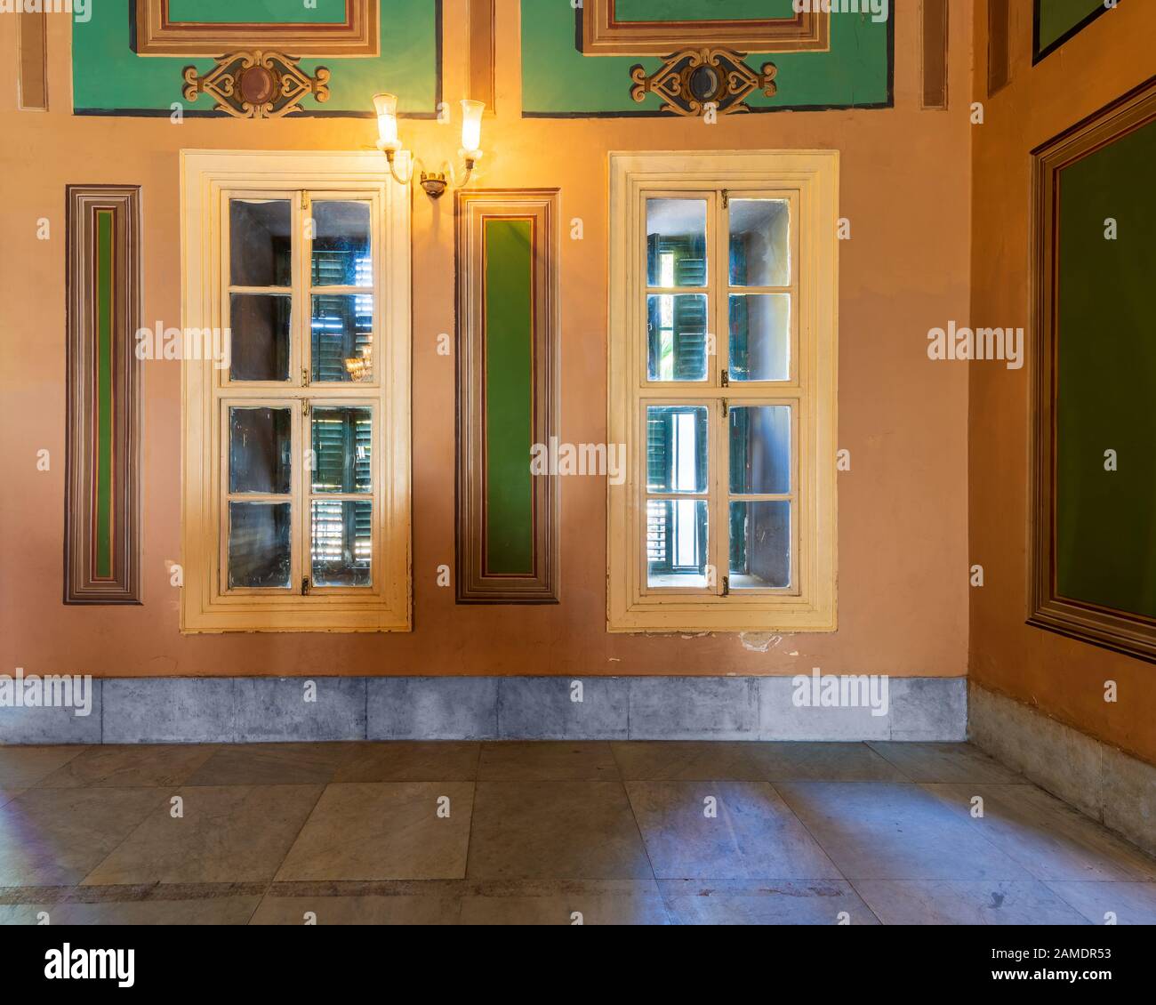 Corner orange wall with two narrow wooden window with closed green shutters, beautiful elegant rectangular green frames, and white tiled marble floor Stock Photo