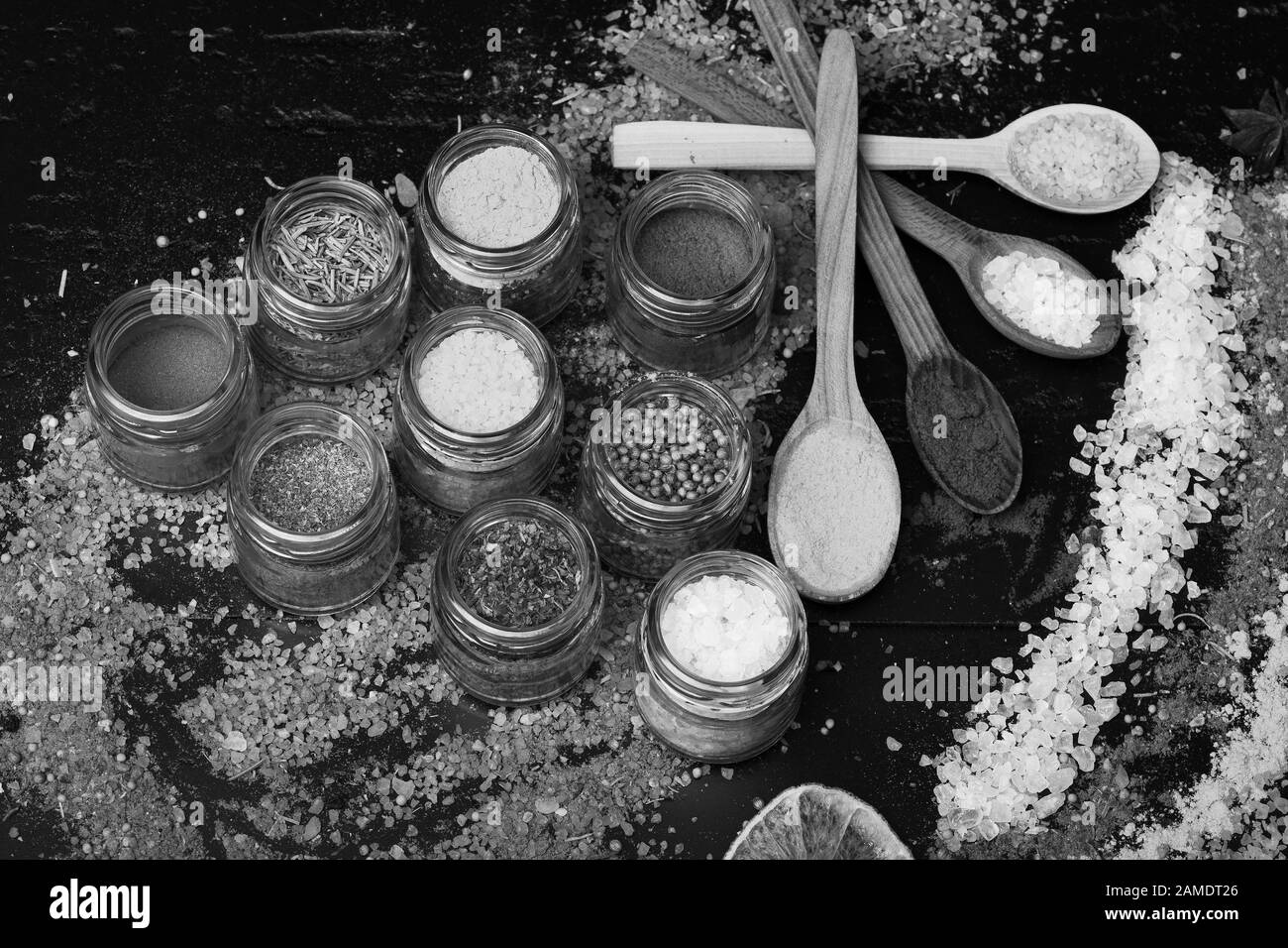 Composition of colored salt and spices near jars with herbs. Wood spoons with paprika, turmeric and sea salt near spice mix scattered on table. Set of spices on dark grey background. Food art concept. Stock Photo