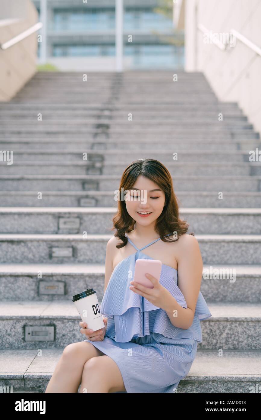 Image of smiling nice woman using cellphone and drinking coffee takeaway while sitting on stairs outdoors Stock Photo