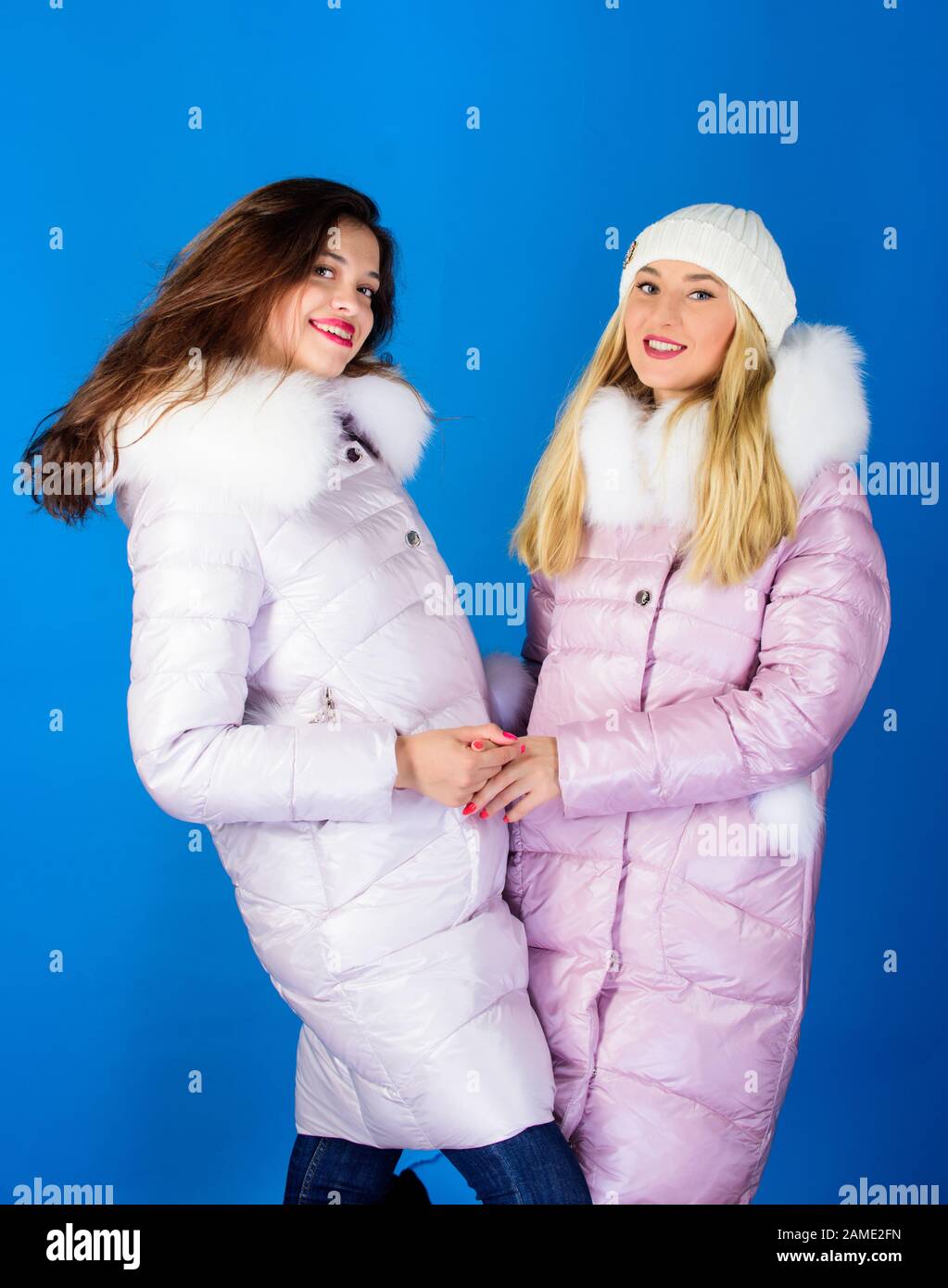 Fashion friends. Winter clothes. Women wear down jacket with furry hood.  Girls smiling makeup faces wear winter jackets blue background. Winter  season. Soft fur. For those wishing stay modern - Stock Image 