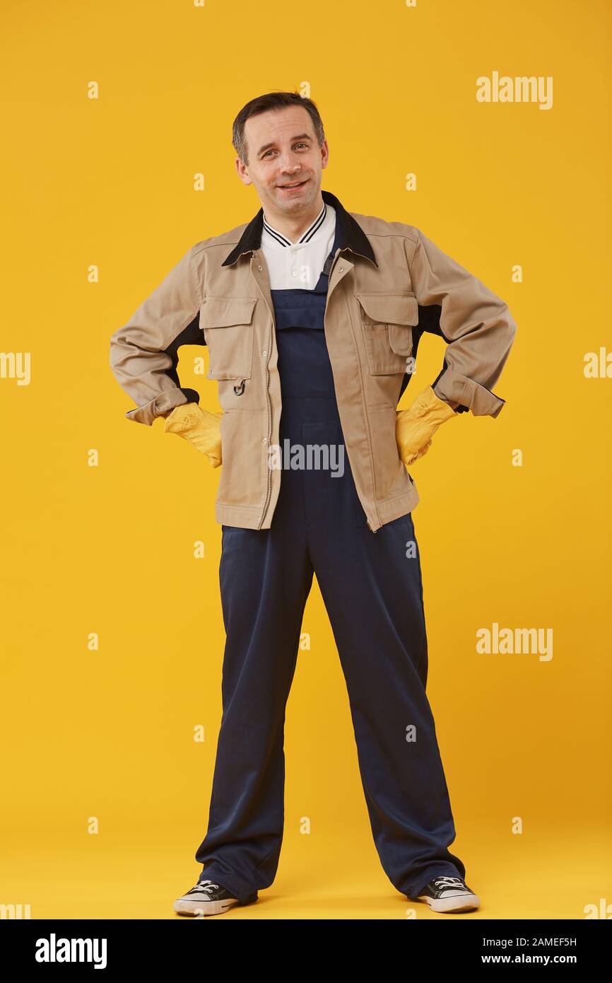 Full length portrait of cheerful mature worker standing against yellow background and posing confidently with hands on hips Stock Photo
