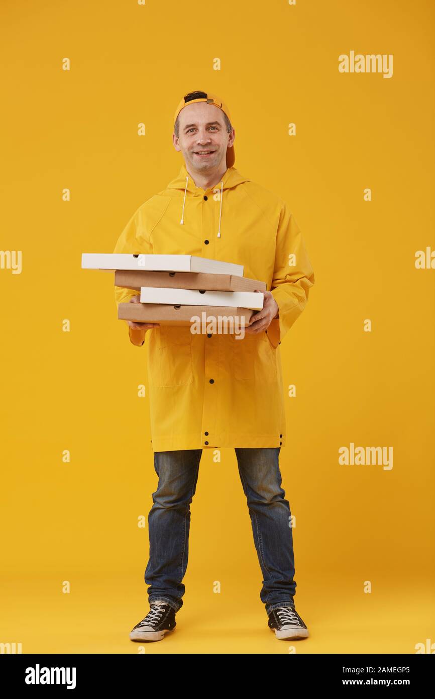 Full length portrait of adult delivery man holding pizza boxes and smiling cheerfully at camera standing against yellow background Stock Photo