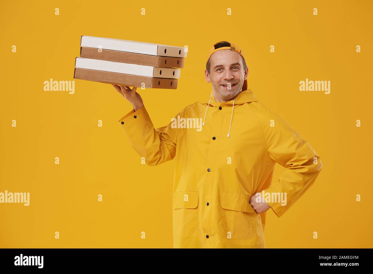 Waist up portrait of cheerful delivery man holding pizza boxes high on one hand and smiling at camera while posing against yellow background, copy space Stock Photo