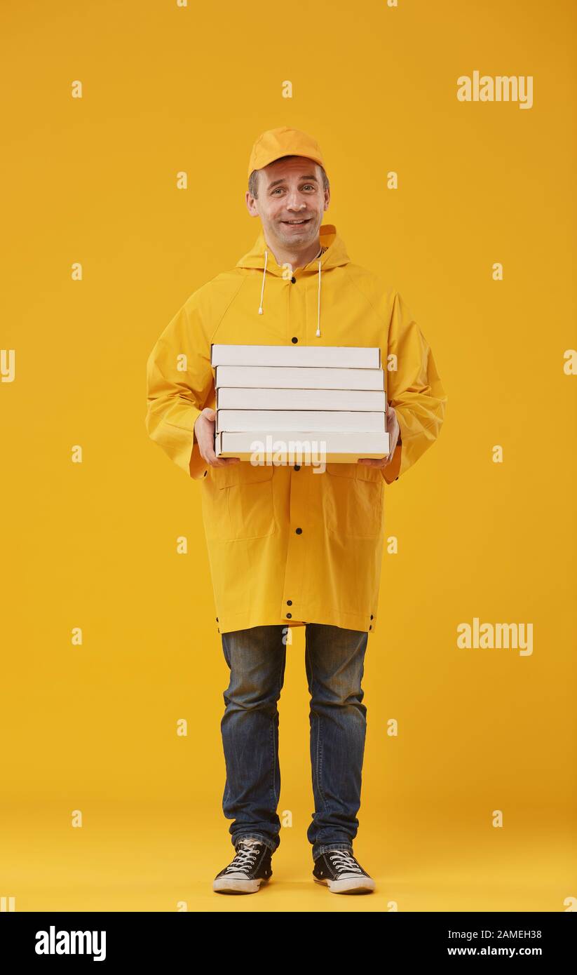 Full length portrait of adult delivery man holding pizza boxes and smiling at camera standing against yellow background Stock Photo