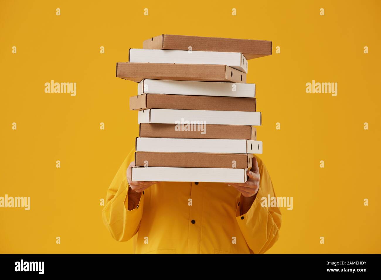 Waist up portrait of unrecognizable delivery man hiding behind pizza boxes while standing against pop yellow background in studio Stock Photo