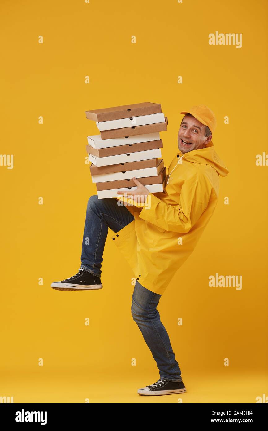 Full length portrait of animated delivery man holding stack of pizza boxes and smiling cheerfully at camera against pop yellow background Stock Photo