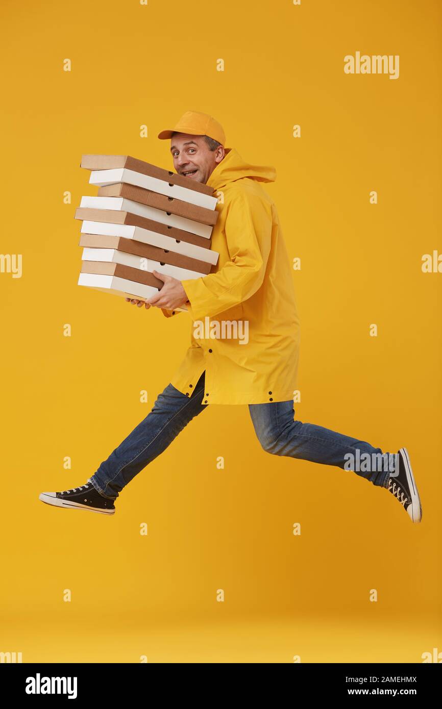 Side view full length of joyful delivery guy jumping high holding pizza boxes against pop yellow background, super fast food service, copy space Stock Photo