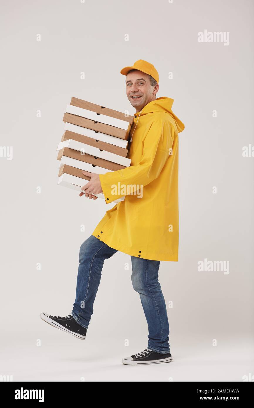 Side view full length portrait of adult delivery man holding stack of pizza boxes and smiling at camera walking against white background Stock Photo