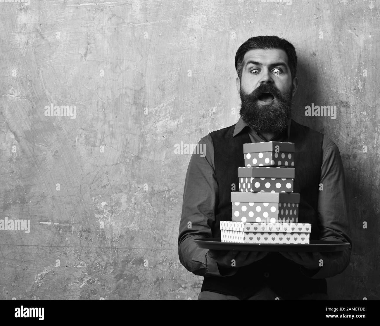 Businessman with shocked face brings gifts for Christmas or birthday. Waiter with pink and red presents on tray. Man with beard holds boxes on beige wall background. Service and shopping concept. Stock Photo