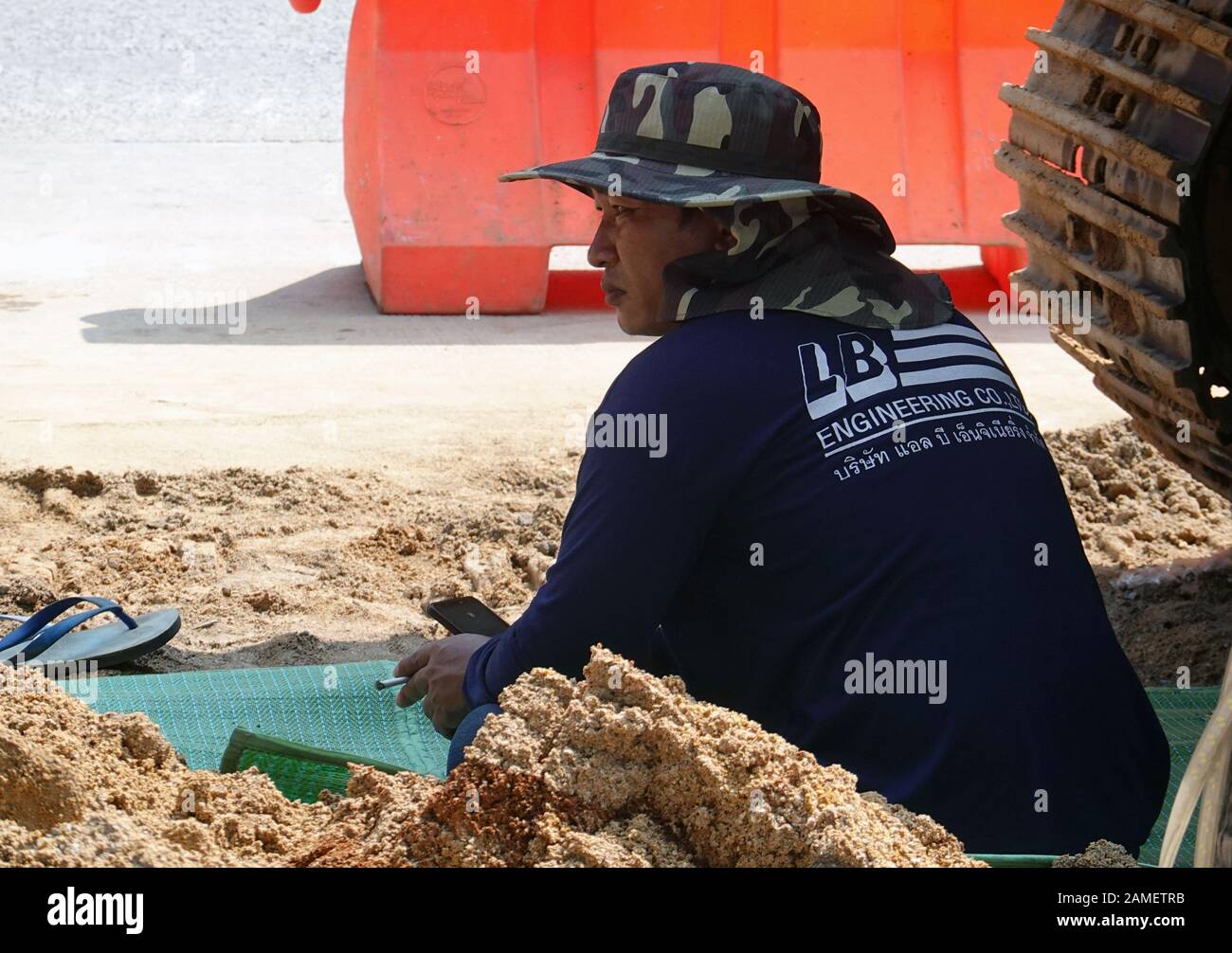 Construction site worker taking a smoke break. The shade of the excavator gives protection from the sun. Stock Photo