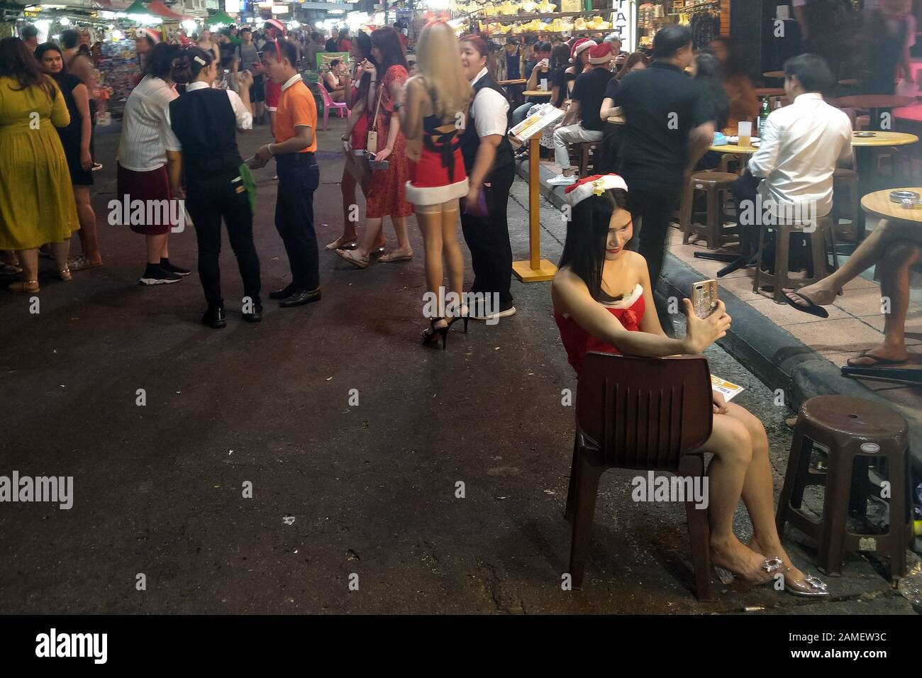 Busy street with lots of people walking and enjoying nightlife. Thai ladyboy with Christmas outfit sitting on chair and taking a selfie. Stock Photo