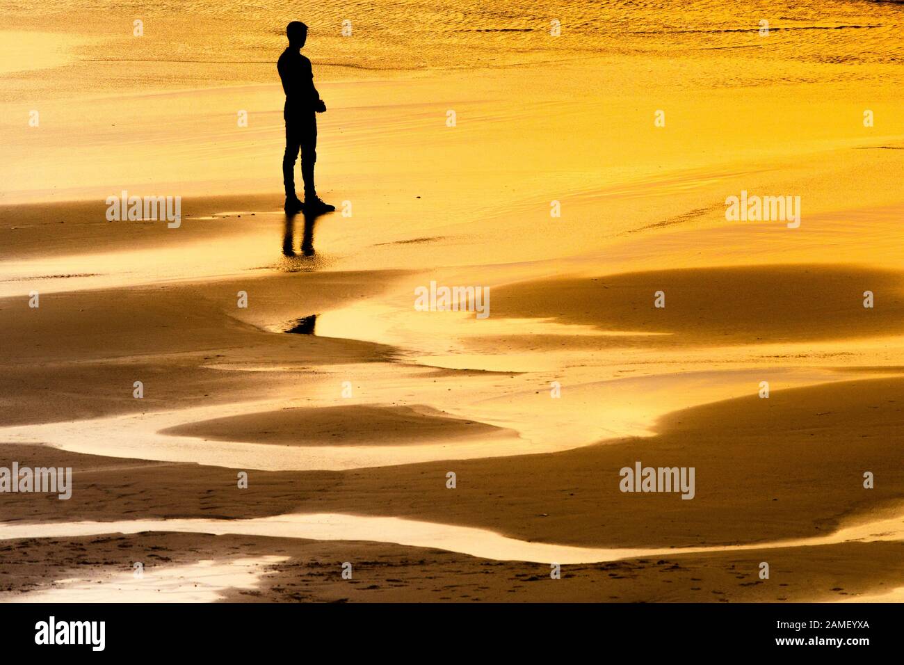 A lone male figure silhouetted by the intense light of the setting sun as he stands looking out to sea at Fistral beach in Newquay in Cornwall. Stock Photo