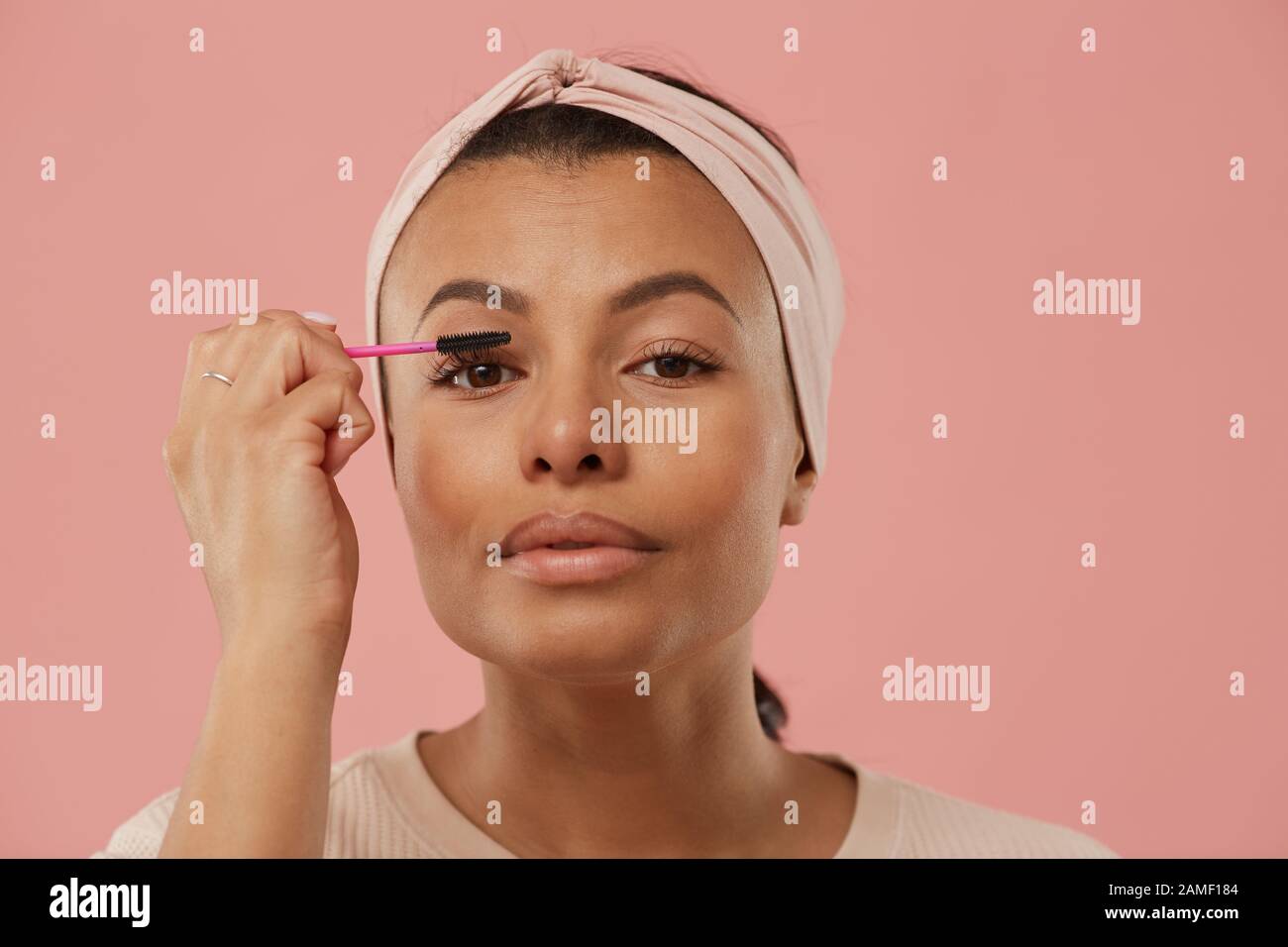 Head and shoulders portrait of beautiful mixed race woman applying mascara while putting on makeup in morning standing against dust pink background, copy space Stock Photo