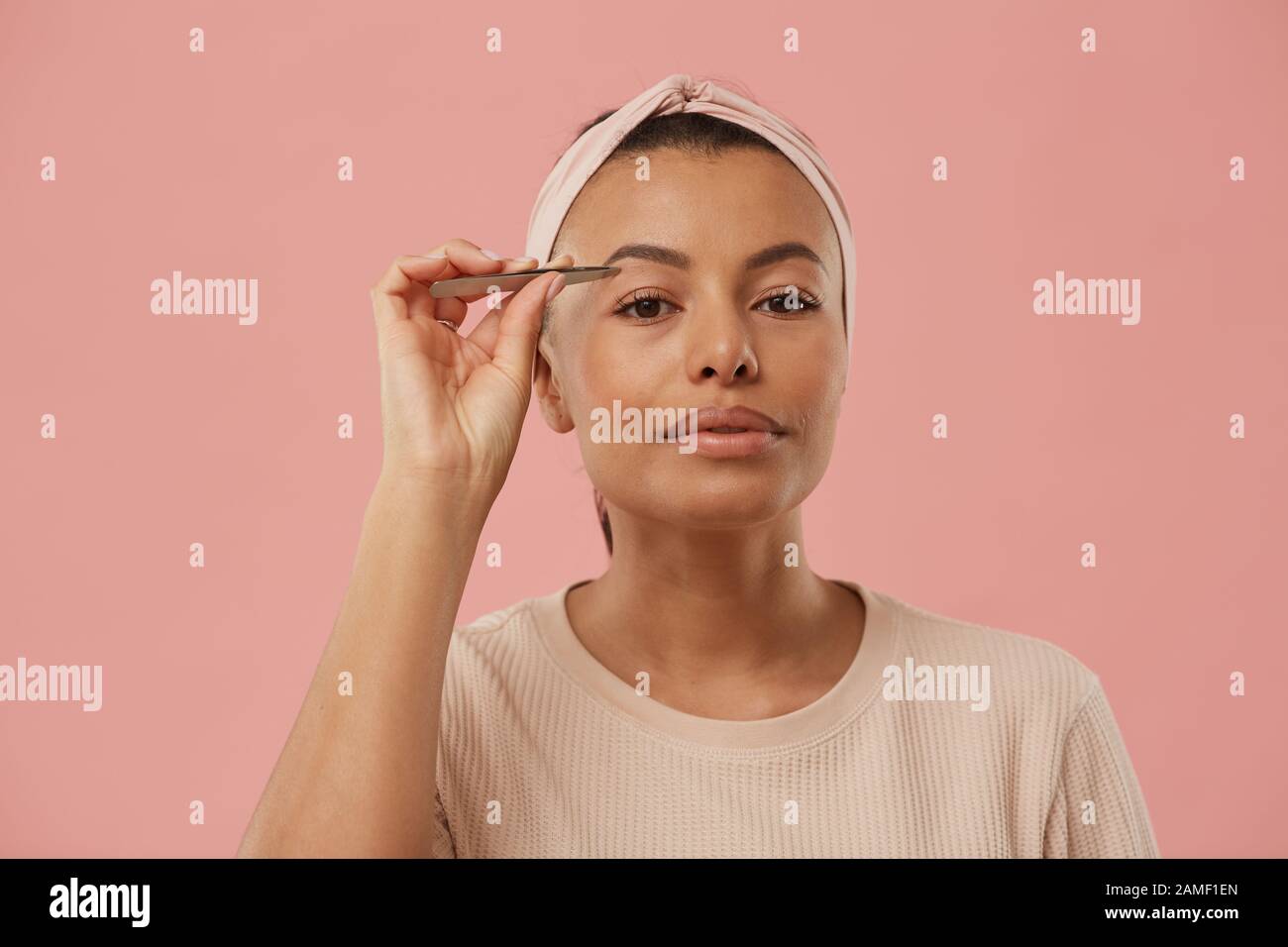 Head and shoulders portrait of beautiful mixed race woman plucking eyebrows while putting on makeup in morning standing against dust pink background, copy space Stock Photo