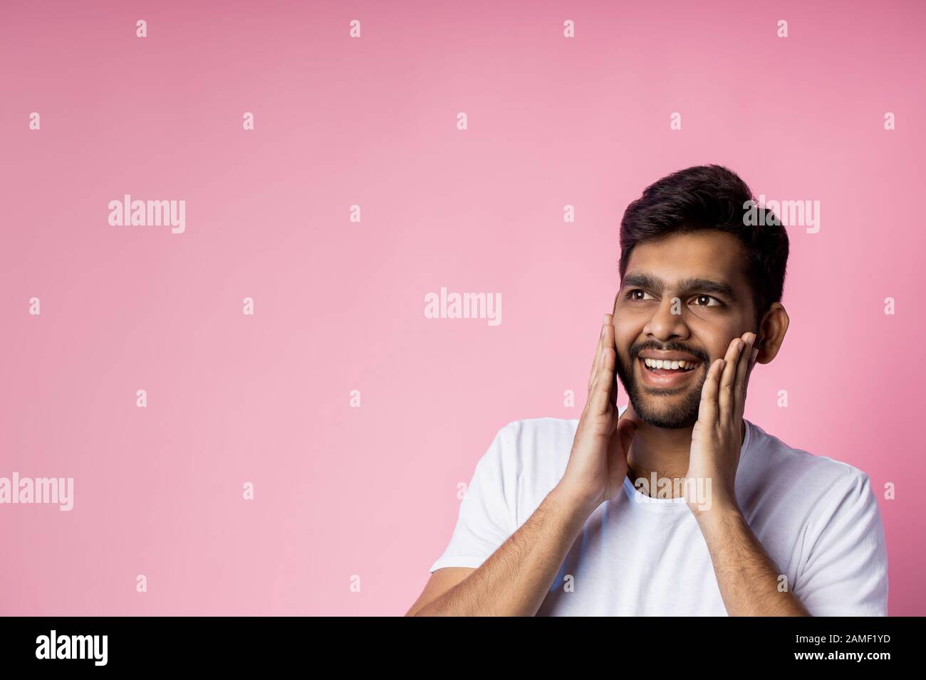 Portrait of pleasantly surprised happy man in white t shirt, touching cheeks with palms, looking aside with amazed expression, isolated on pink backgr Stock Photo