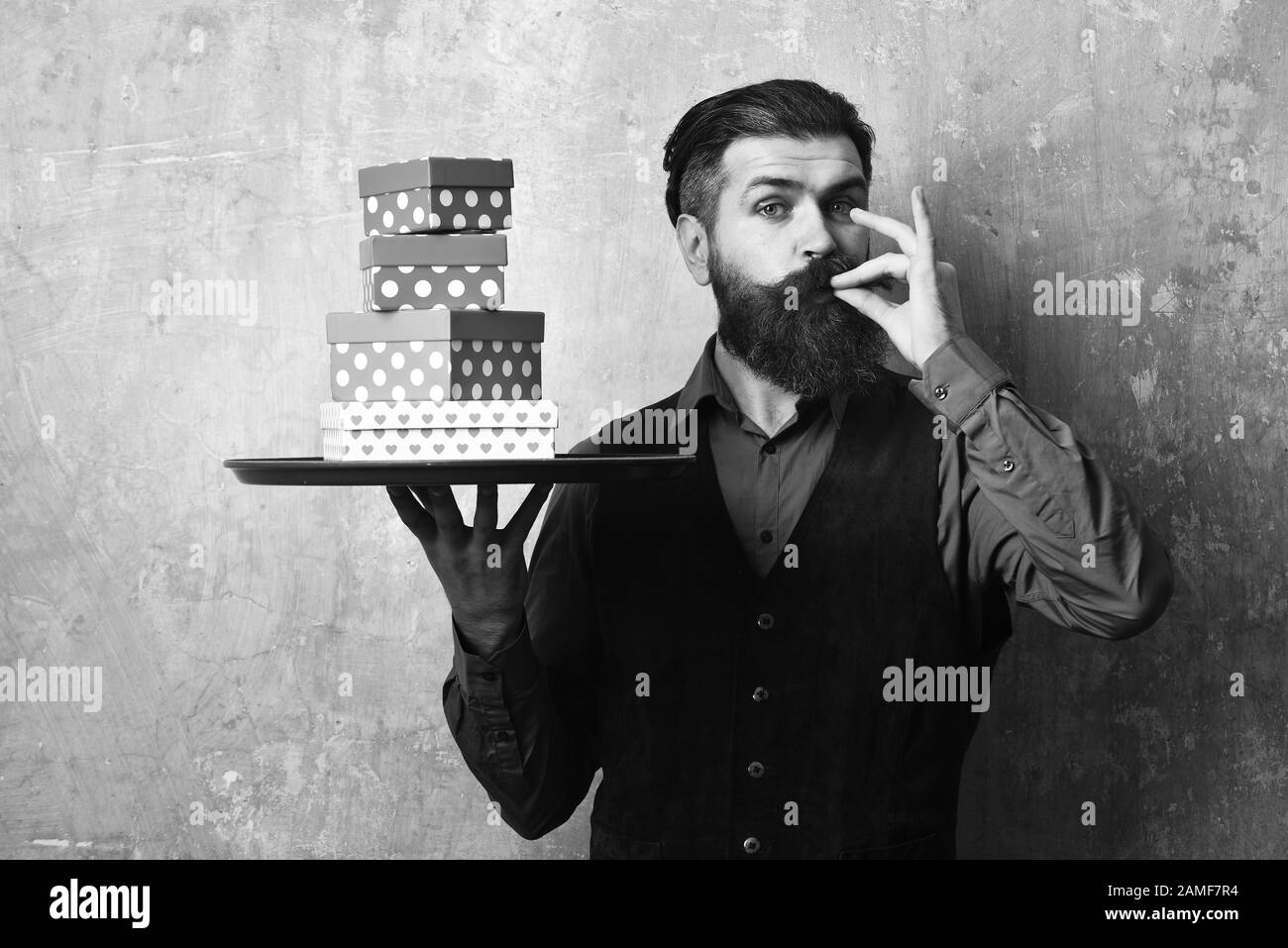 Waiter with presents on tray. Businessman with satisfied face brings gifts for Christmas. Service and purchase concept. Man with beard shows perfect taste sign holding boxes on beige wall background. Stock Photo
