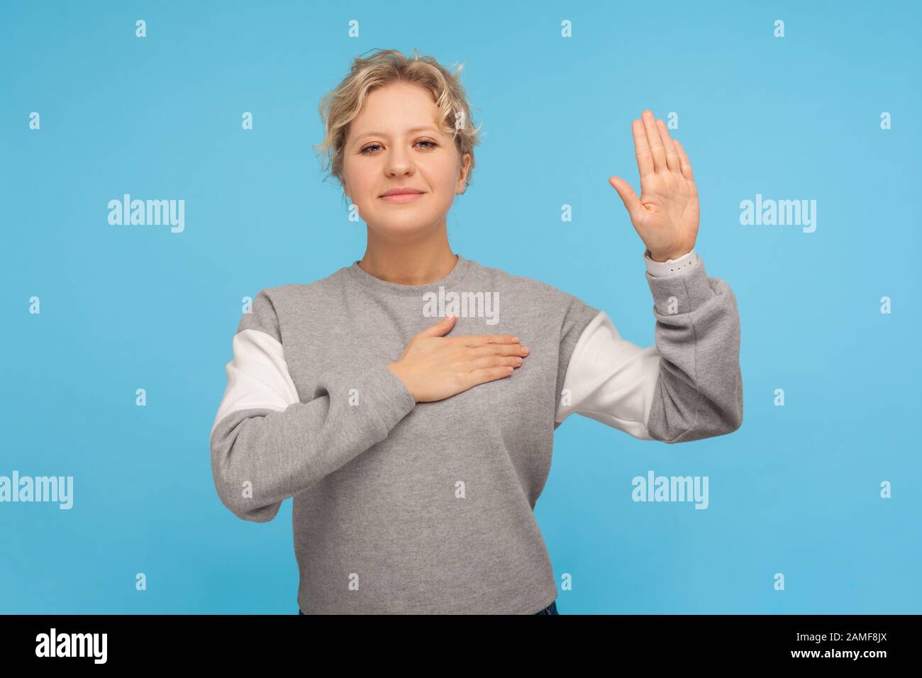 Portrait of patriotic citizen, honest adult woman with curly hair in sweatshirt keeping hand on chest and raising palm, making loyalty promise, oath. Stock Photo