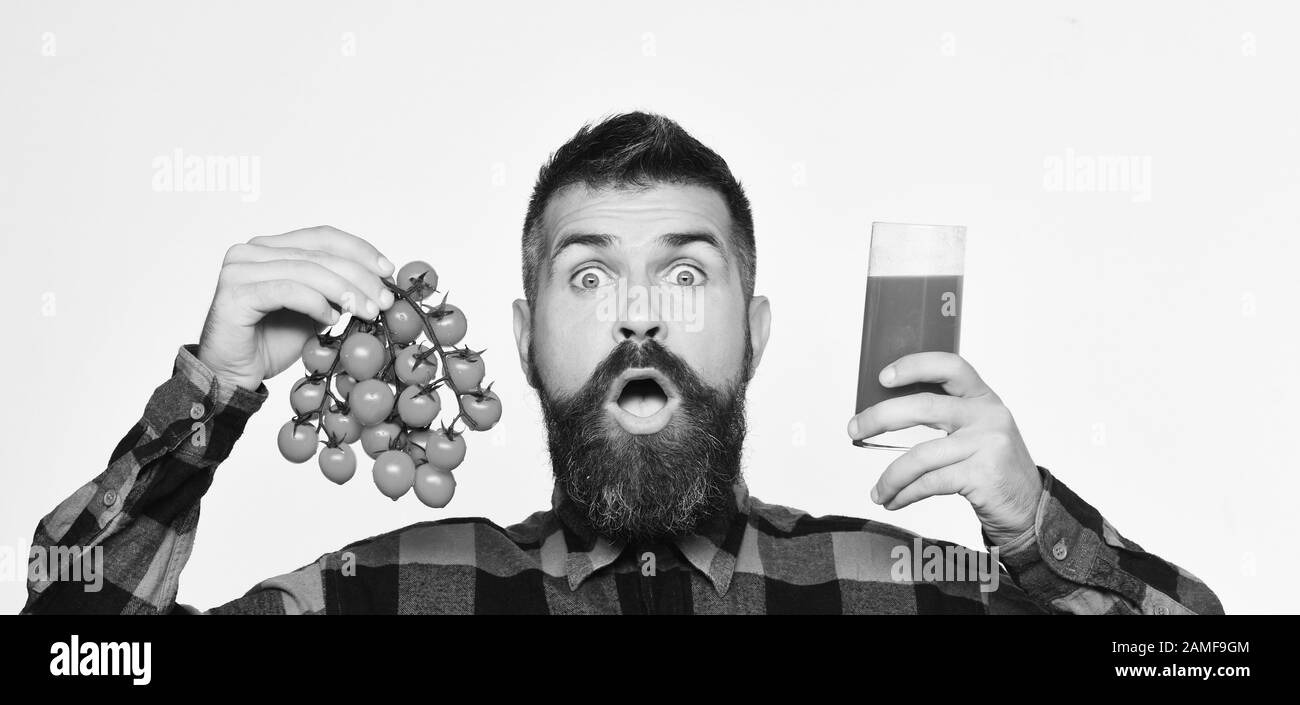 Man with beard holds glass of juice isolated on white background. Farmer with surprised face shows tomato juice and bunch of cherry tomatoes. Guy holds homegrown harvest. Farming and autumn concept. Stock Photo