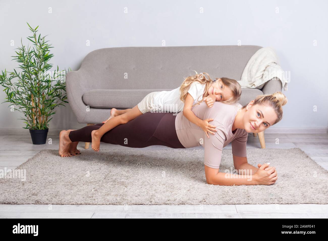 Cheerful mother doing plank workout with little daughter on her back, having fun smiling at camera, happy family doing gymnastics exercise together, f Stock Photo