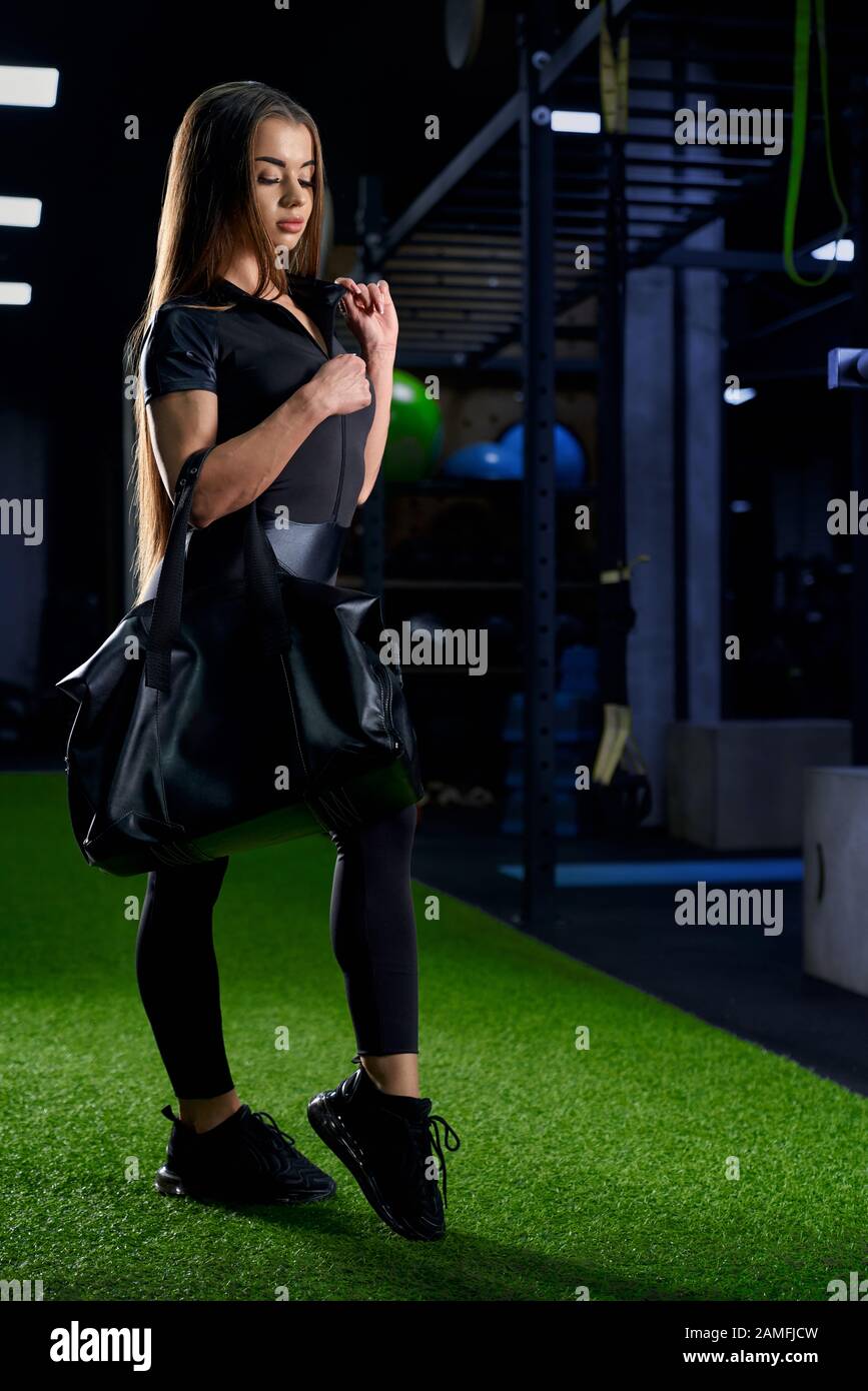 Side view of young attractive fit woman posing with sport bag in gym on fake grass. Pretty female with long hair in black sportswear going home after hard training. Workout, fitness concept. Stock Photo
