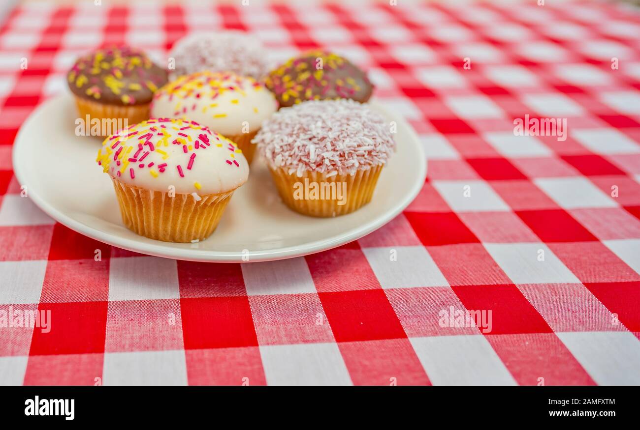 8 Selective focus on a cup cake on a plate of six cupcakes on a plain white plate on a red and white checked table cloth Stock Photo