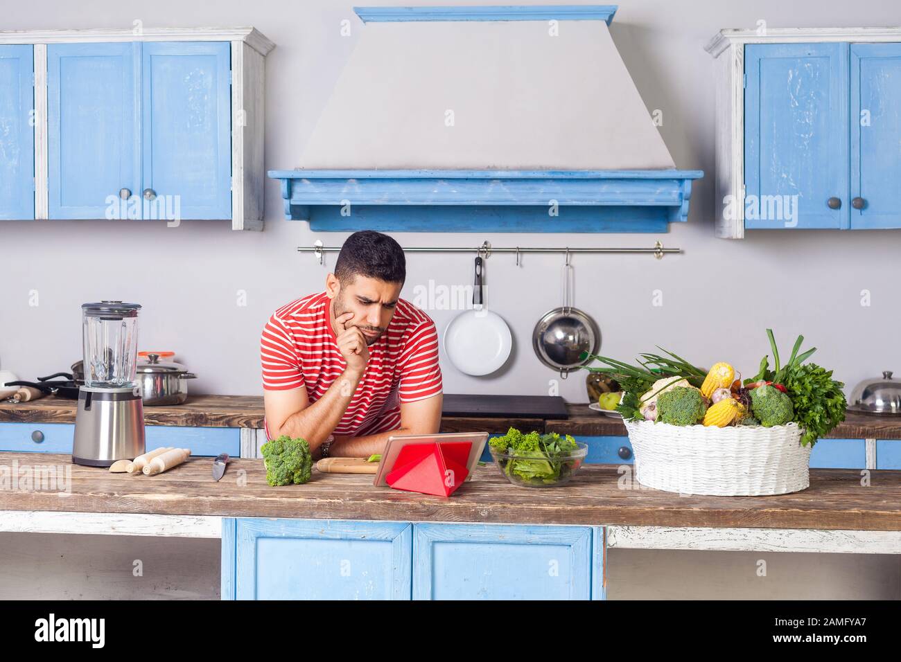 Thoughtful confused man watching food blog using tablet in modern kitchen, looking for dish recipe on internet to cook vegetarian salad with green fre Stock Photo
