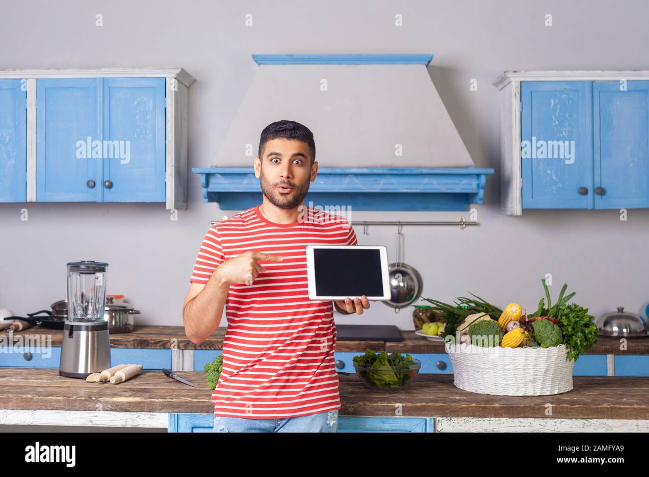 Surprised young man standing in kitchen and pointing at tablet, looking at camera with amazement, recommending food recipes from internet, unbelievabl Stock Photo