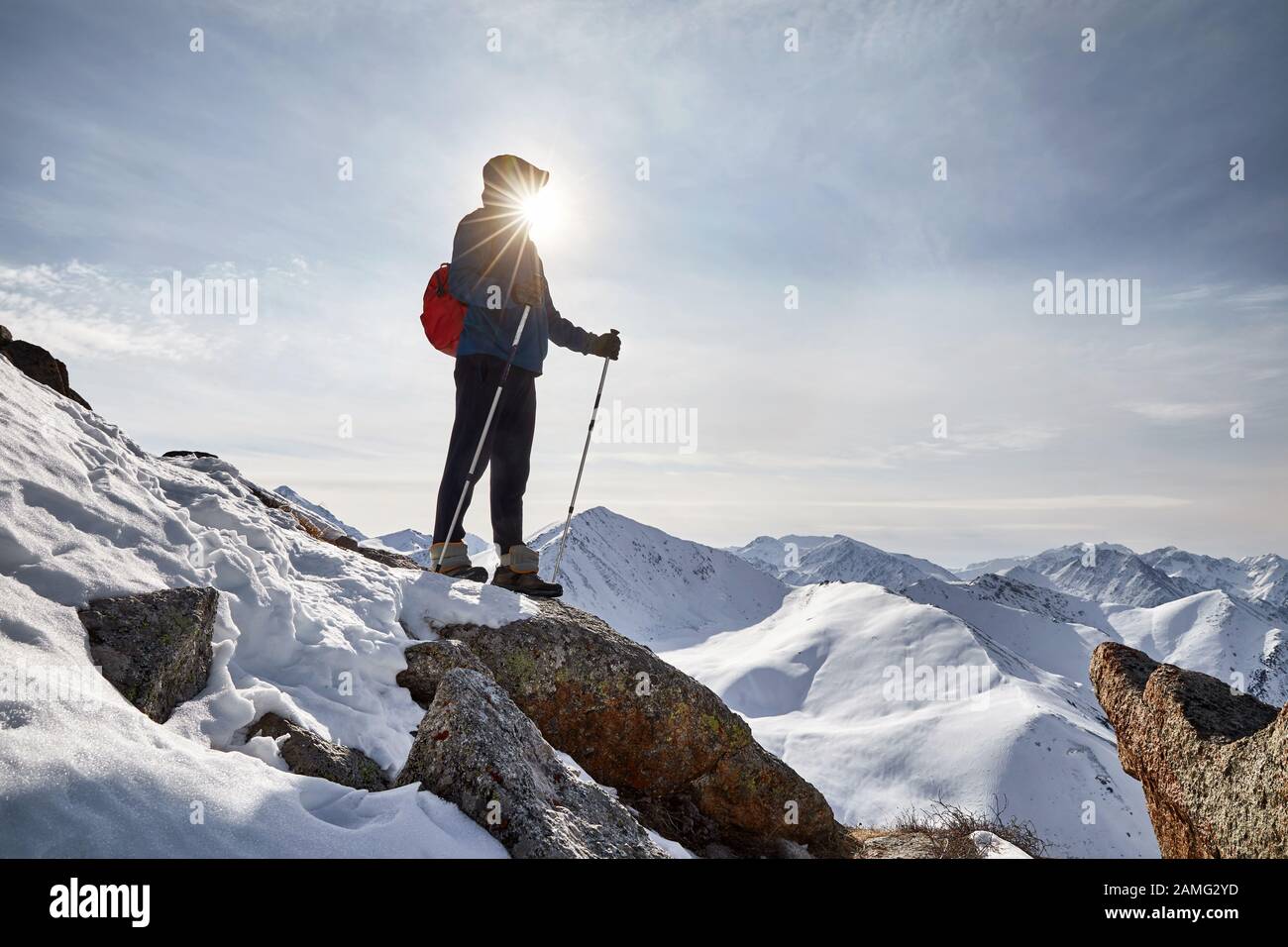 Climber in blue jacket with trekking poles is standing on the rock against the sun at high snowy mountains. Outdoor climbing and mountaineering concep Stock Photo