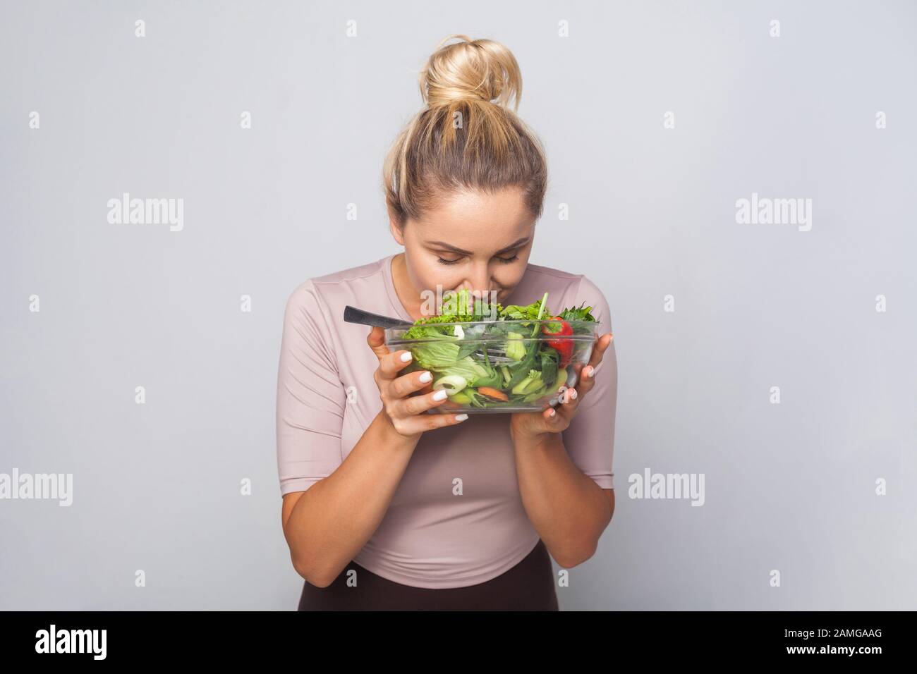 Portrait of young woman with hair bun sniffing bawl of green vegetable salad, enjoying smell of fresh healthy food, vegetarian diet, vegan nutrition. Stock Photo