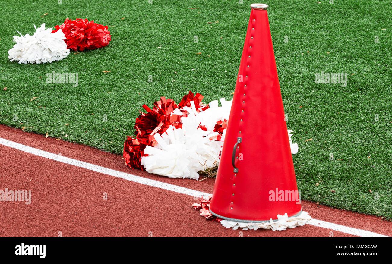 A red megaphone used by cheerleaders on the track next to red and white pom poms on the green turf. Stock Photo