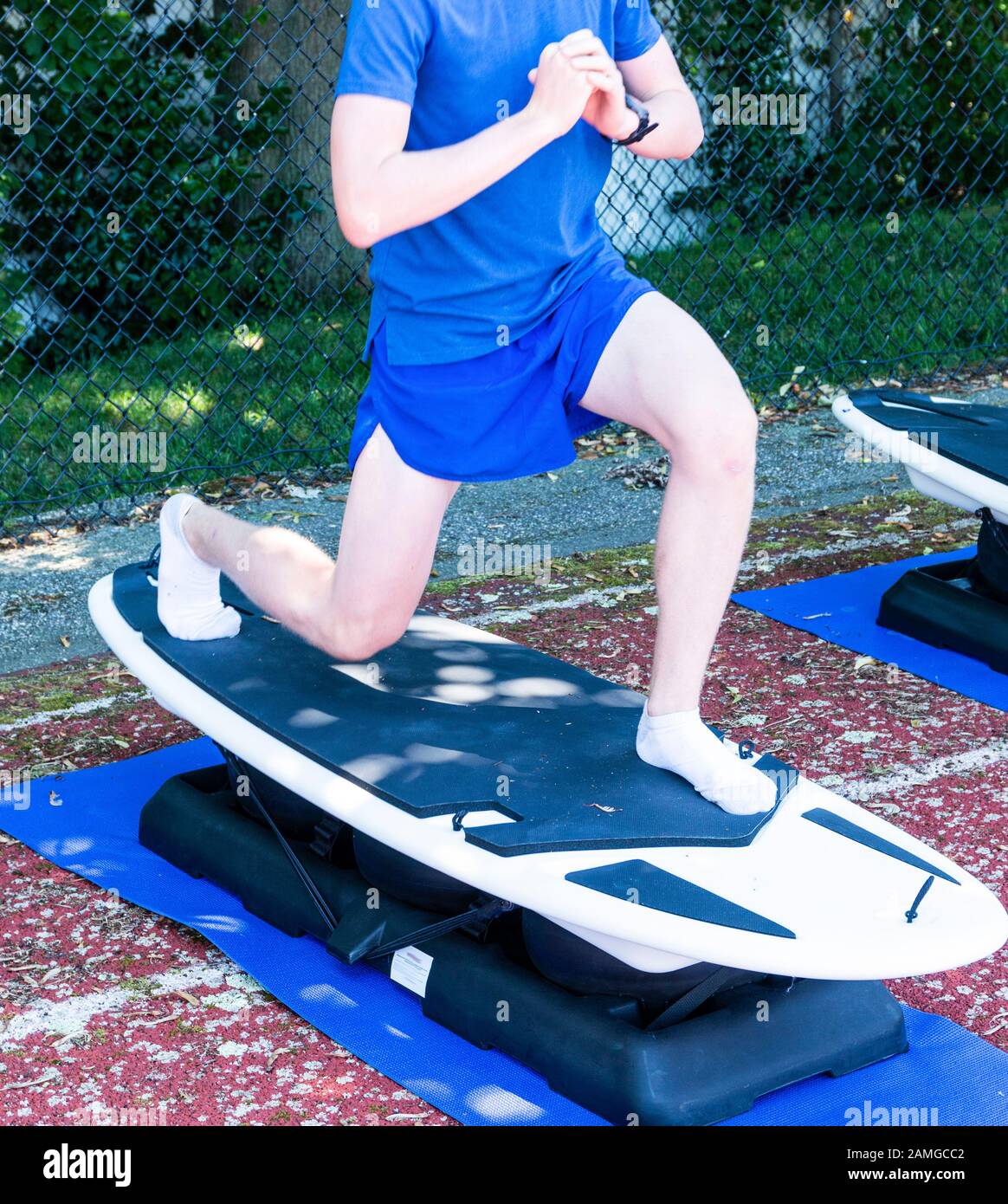 A teenage runner performing lunges on a unstable surboard machine for strength and stability practice during a summer camp in the shade on a track. Stock Photo