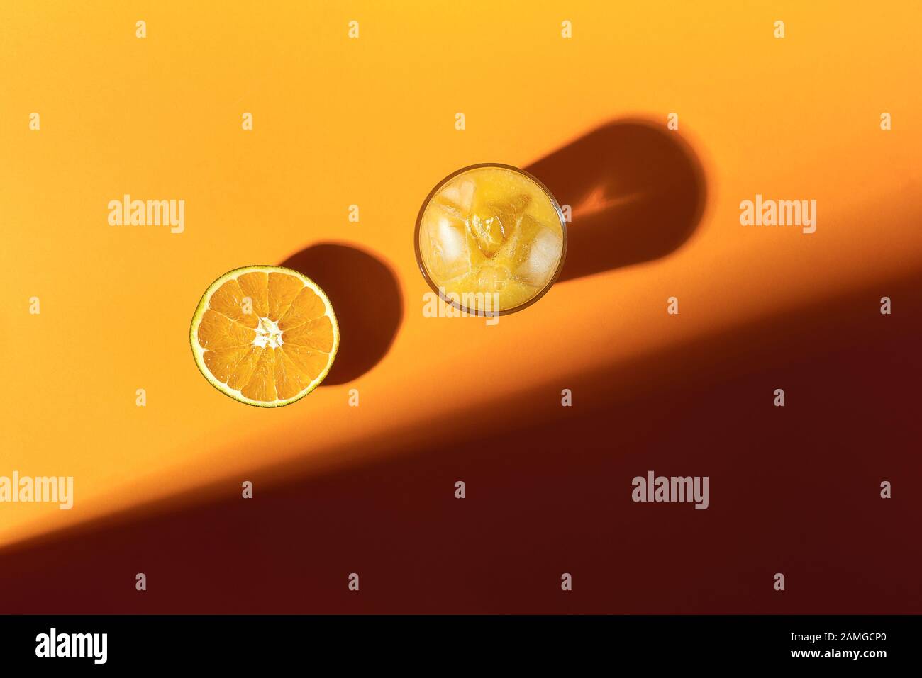 Fresh orange juice glass with ice and half of orange fruit, in sunlight. Above view of citrus fruit and summer refreshment on orange background. Stock Photo