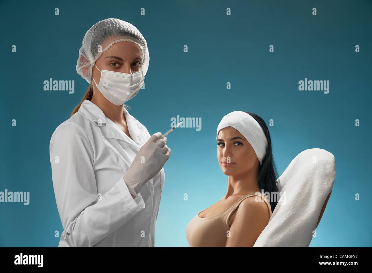 Side view of female cosmetologist in mask and coat holding syringe. Brunette woman patient with towel on head sitting in chair, smiling, looking at camera, isolated on blue. Concept of cosmetology. Stock Photo