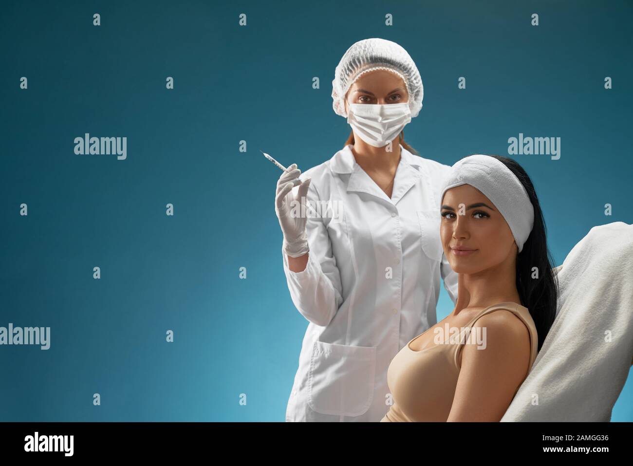 Side view of female cosmetologist in mask and coat holding syringe. Brunette woman patient with towel on head sitting in chair, smiling, looking at camera, isolated on blue. Concept of cosmetology. Stock Photo