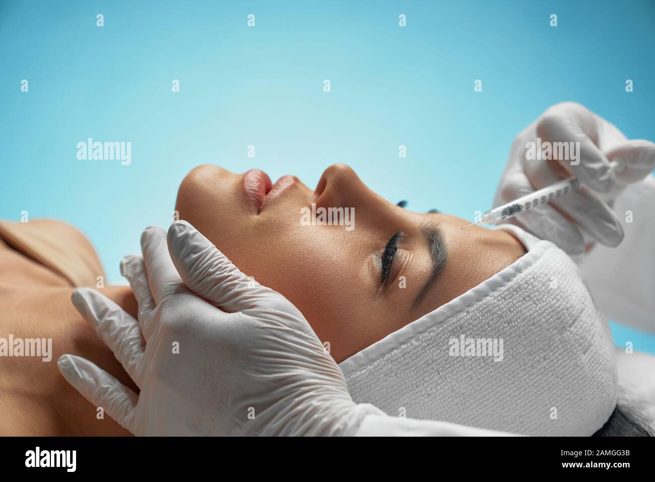 Close up of botox injection in female forehead. Side view of cosmetologist using syringe with special liquid, holding chin while patient with towel on head lying. Concept of cosmetology, beauty. Stock Photo