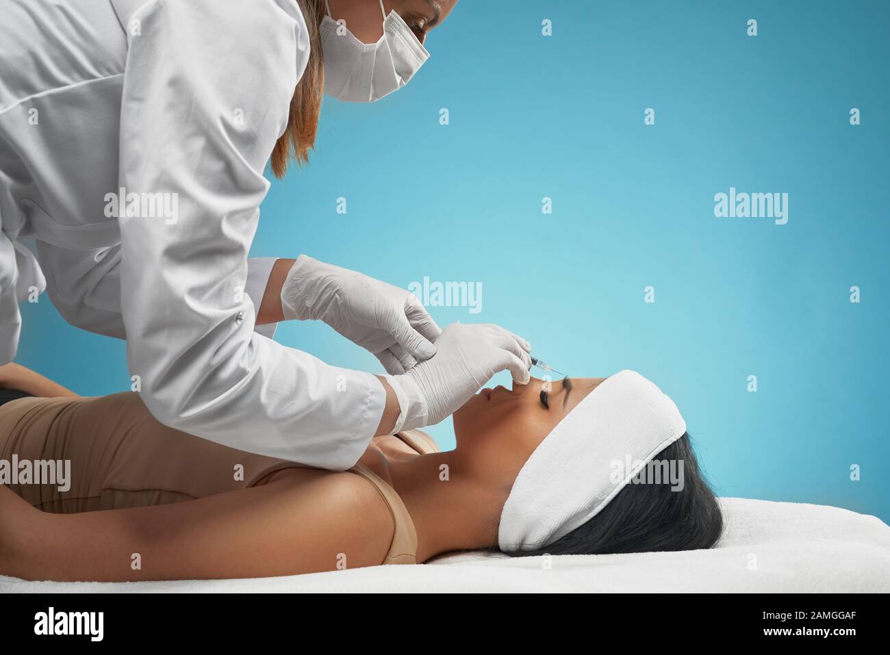 Side view of injection in female forehead. Cosmetologist in doctors coat using syringe with botox and cotton pad, brunette patient with closed eyes lying on couch. Concept of cosmetology, beauty. Stock Photo