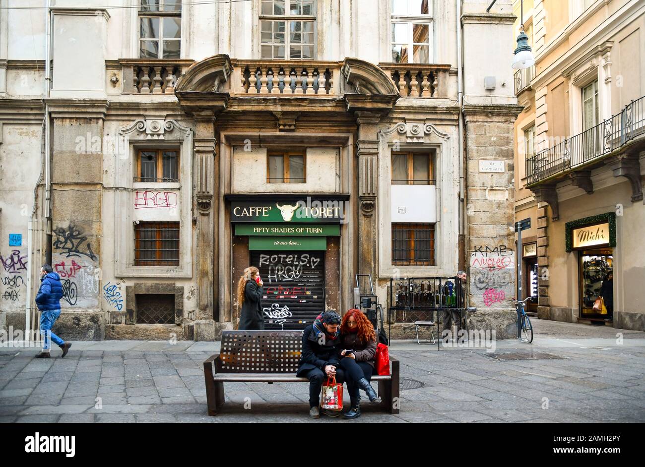 Glimpse of Via Garibaldi street with a couple sitting on a bench and people walking in front of the closed Caffé Toret, Turin, Piedmont, Italy Stock Photo