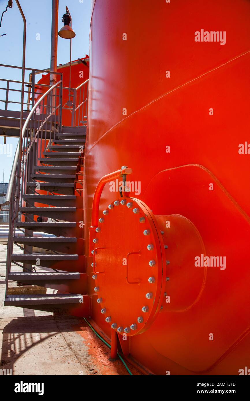 Orange steel storage tanks with sulfuric acid on warehouse of oil refinery plant. The metal stairway and round manhole. Close-up. Stock Photo