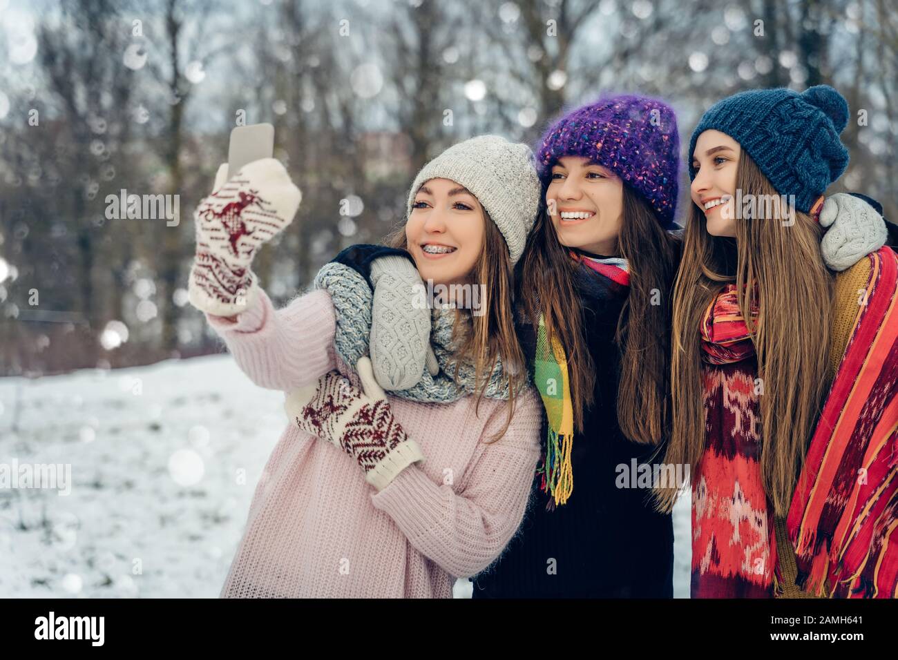 Three women friends outdoors in knitted hats using mobile phone on a snowy cold weather. Group of young female friends enjoying taking selfies Stock Photo