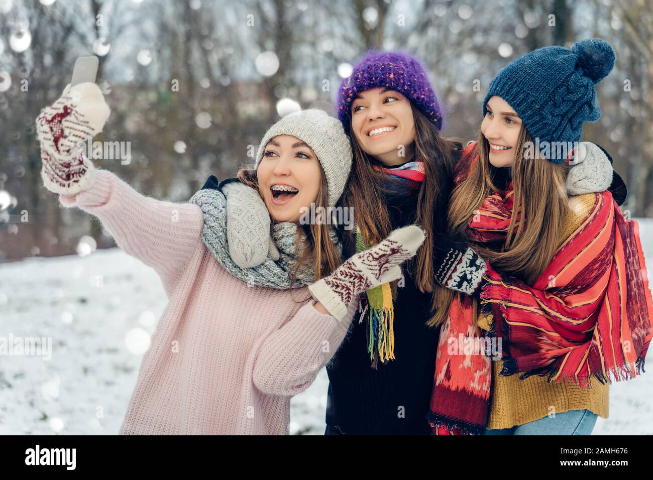 Three women friends outdoors in knitted hats using mobile phone on a snowy cold weather. Group of young female friends enjoying taking selfies Stock Photo