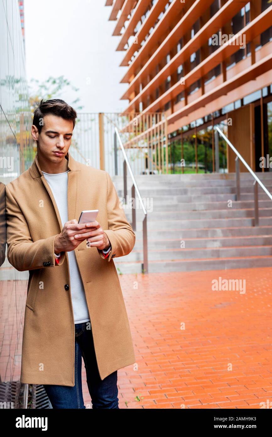 Young man wearing winter clothes in the street using the phone. Young guy  with modern hairstyle with coat, blue jeans and t-shirt Stock Photo - Alamy