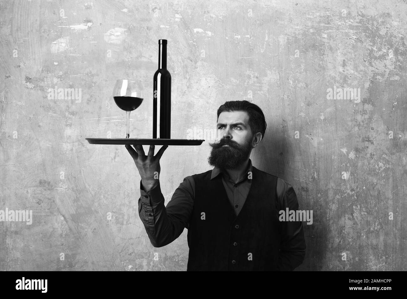 Service and catering concept. Man with beard and mustache holds wine on beige wall background. Waiter looks at glass and bottle of red wine on tray. Barman with curious face serves wine glass. Stock Photo