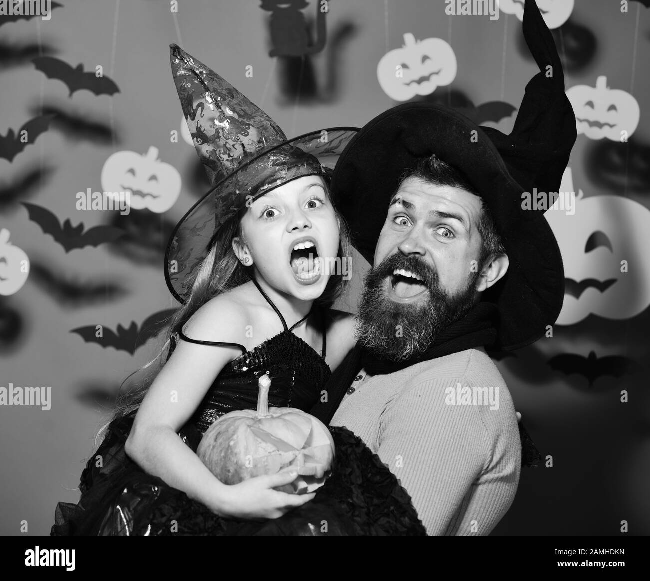 Wizard and little witch in black hats hold pumpkin. Father and daughter in costumes. Girl and bearded man with screaming faces on red background with spooky symbols. Halloween party and decor concept Stock Photo