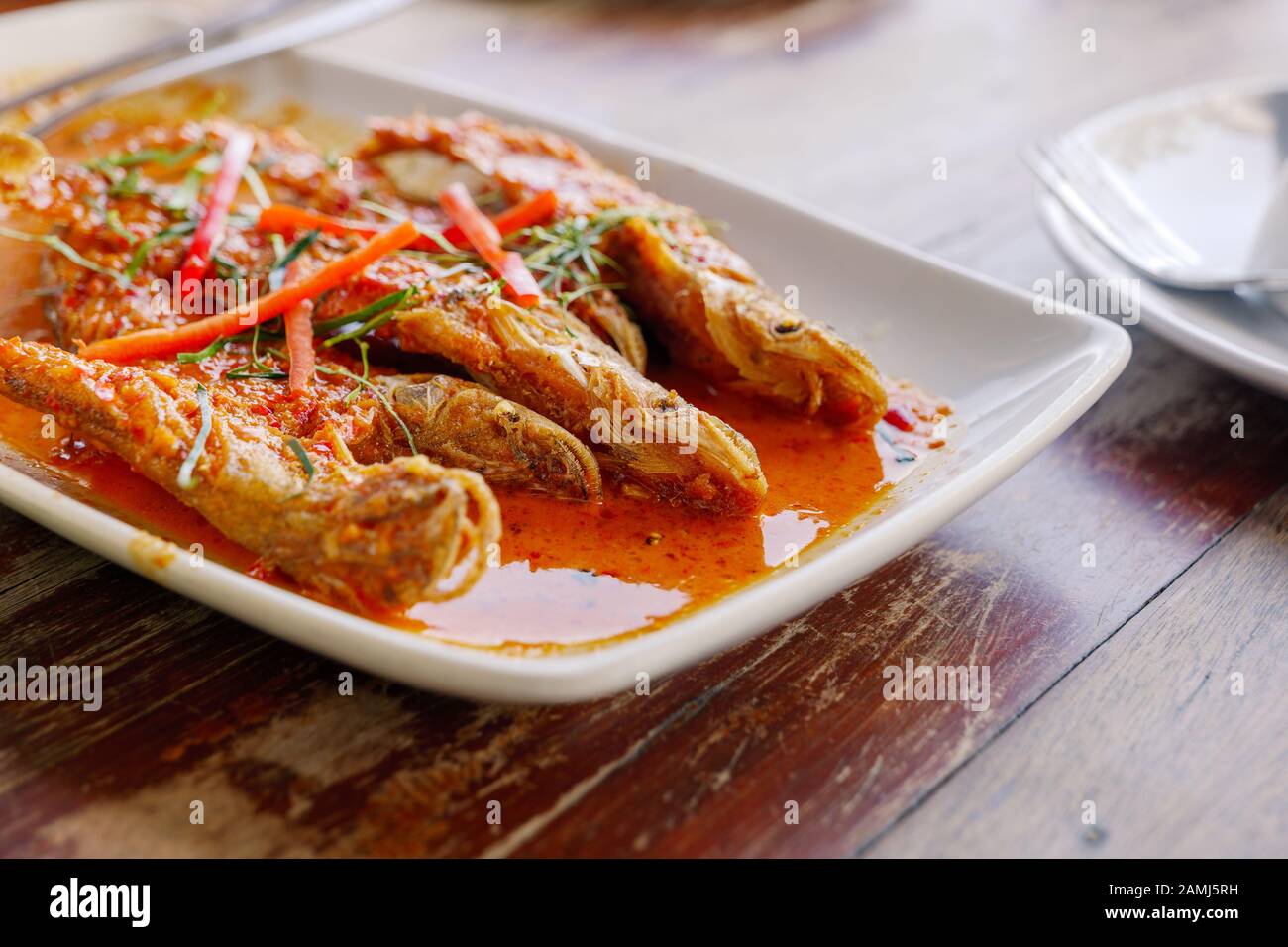 Close up view of Spicy Red Curry with crispy fried soft fish, Sheatfish, on white dish and wooden table. Stock Photo