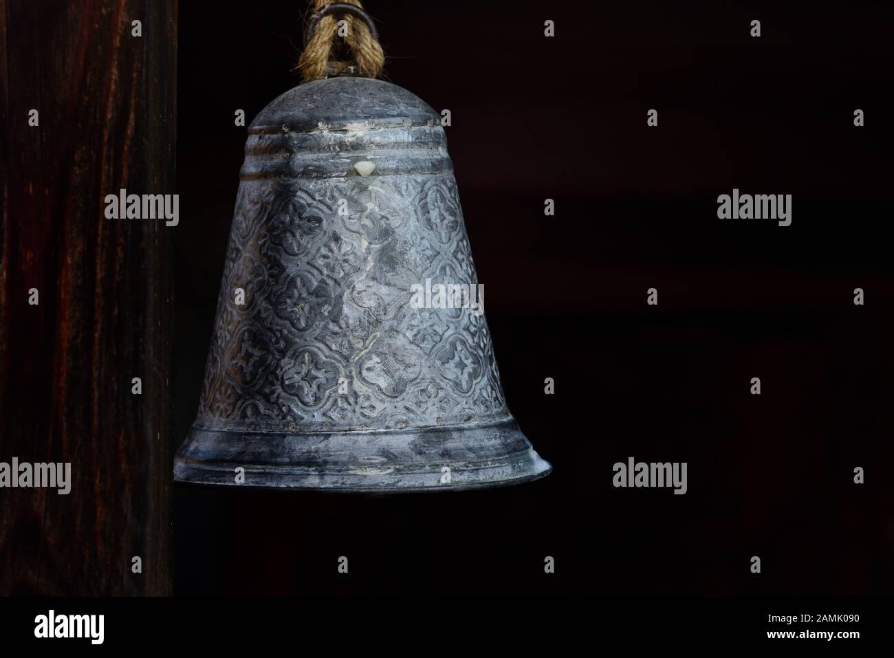 A metal bell hangs rustic on a cord next to a wooden beam against a dark background with space for text Stock Photo