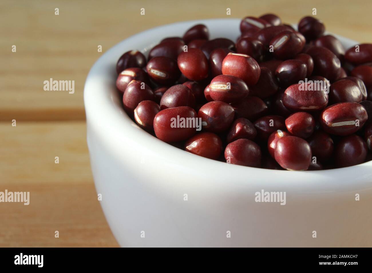 A plain sunlit white bowl of Adzuki beans, also known as red mung bean, in close up, on a plain wooden table. With copyspace. Stock Photo